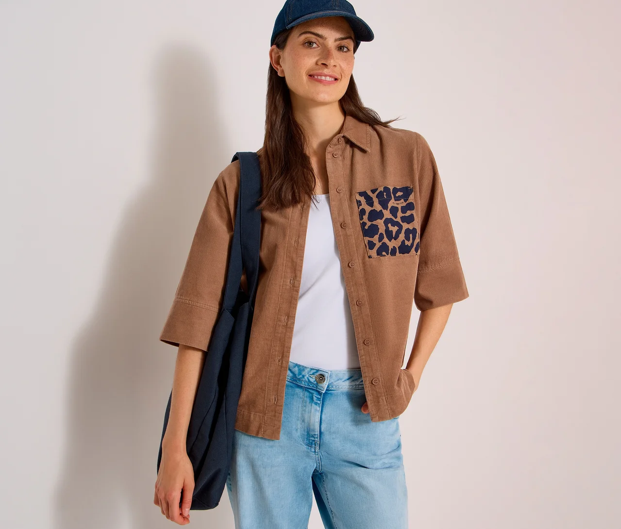 Lächelnde Frau mit dunkelblauer Kappe, trägt eine braune CECIL Hemdbluse mit Leopardenmuster-Tasche, helle Jeans und eine dunkelblaue Tasche.