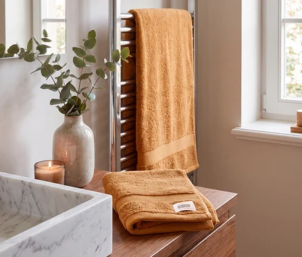 Zwei braune SCHÖNER WOHNEN-Kollektion Cuddly Handtücher in einem Badezimmer. Eines hängt an einem verchromten Handtuchwärmer, ein weiteres liegt gefaltet auf einem Holzwaschtisch neben einem Marmorwaschbecken, einer brennenden Kerze und Eukalyptus in einer Vase.