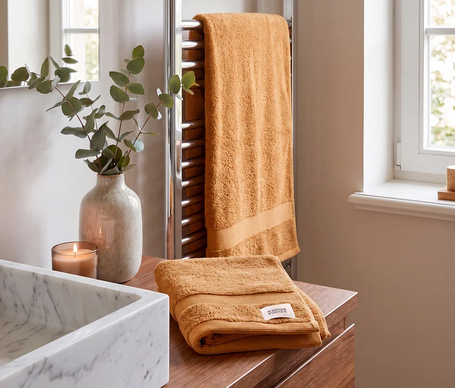 Zwei braune SCHÖNER WOHNEN-Kollektion Cuddly Handtücher in einem Badezimmer. Eines hängt an einem verchromten Handtuchwärmer, ein weiteres liegt gefaltet auf einem Holzwaschtisch neben einem Marmorwaschbecken, einer brennenden Kerze und Eukalyptus in einer Vase.