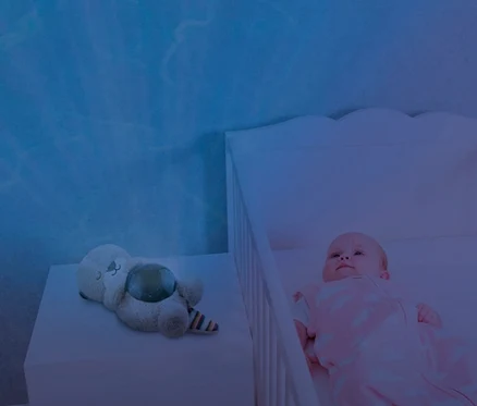 Baby liegt im Gitterbett neben ZAZU-Ozeanprojektor »Otto«.