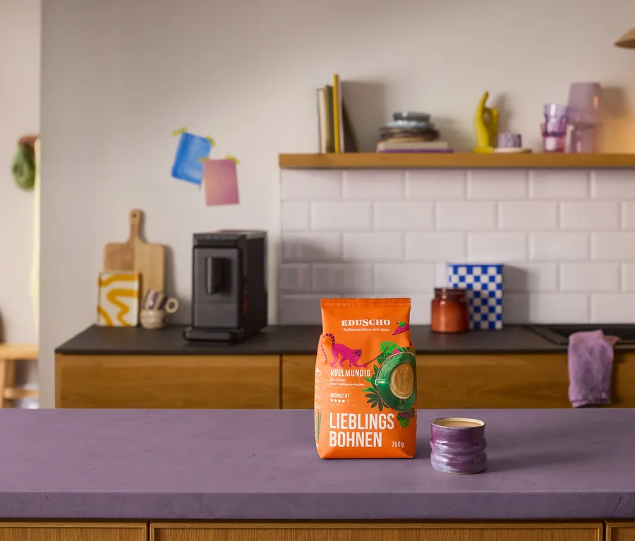 Ein orangefarbener Beutel Eduscho Lieblingsbohnen Vollmundig und eine lila Tasse stehen auf einer lila Arbeitsplatte. Im Hintergrund ist ein schwarzer »Esperto mini« Kaffeevollautomat auf einer dunklen Küchenzeile mit Holzschränken und weißem Fliesenspiegel zu sehen.