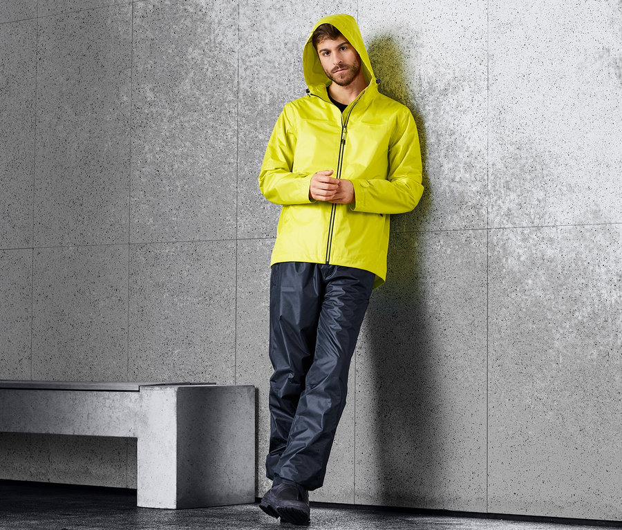 Mann lehnt an einer Wand und trägt eine gelbe Jacke und eine dunkelblaue Unisex-Regenhose.
