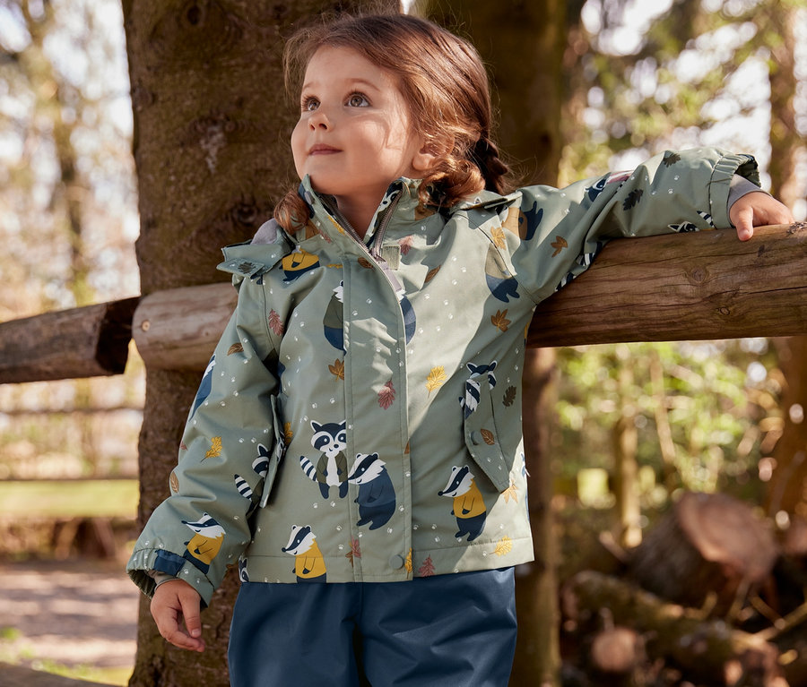 Ein Mädchen steht an einem Holzzaun und trägt Gummistiefel, eine dunkelblaue Thermo-Regenhose und eine Thermo-Regenjacke mit Dachsprint.