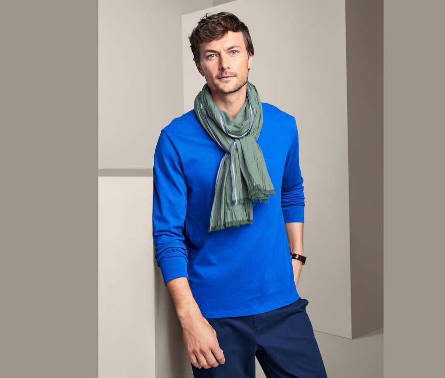 Mann mit braunen Haaren und blauen Augen trägt ein blaues T-Shirt und einen Webschal.