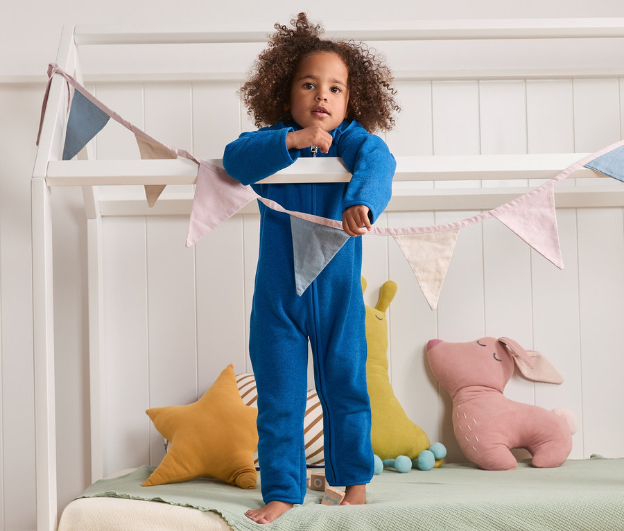 Kind steht in blauem Strickfleece-Einteiler mit Kapuze mit recyceltem Material auf einem weißen Bett mit dekorativer Wimpelgirlande.