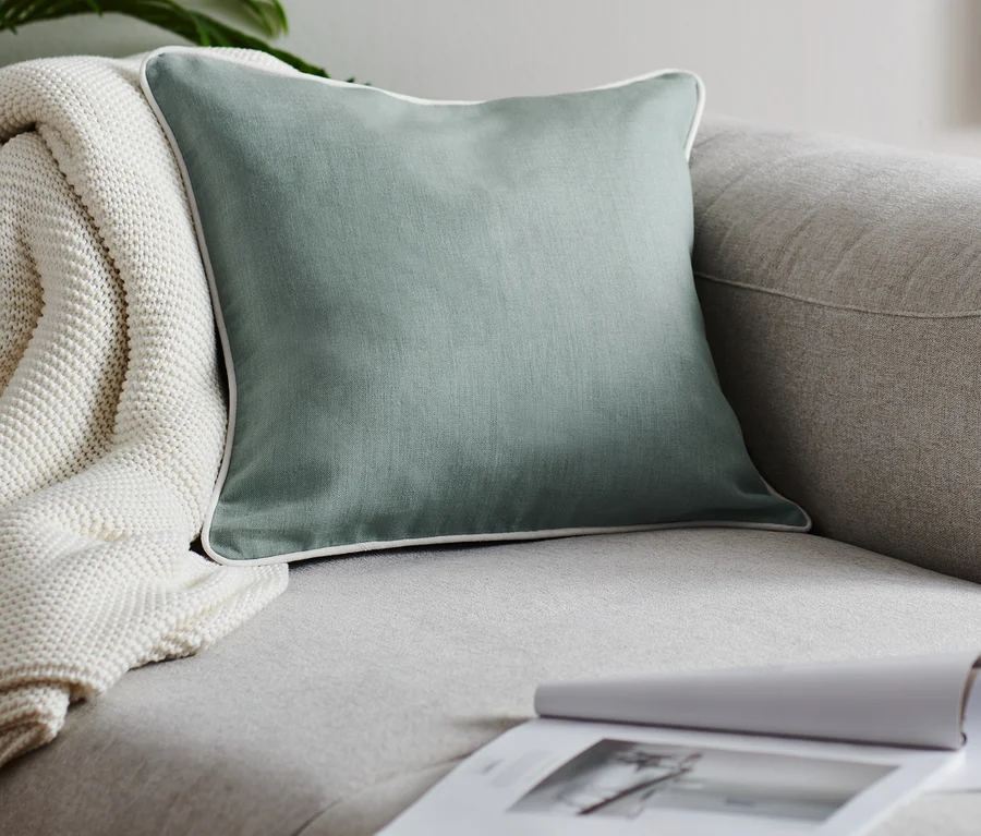 Ein grüner Leinen-Dekokissenbezug liegt auf einem grauen Sofa neben einer weissen Strickdecke und einem offenen Buch.