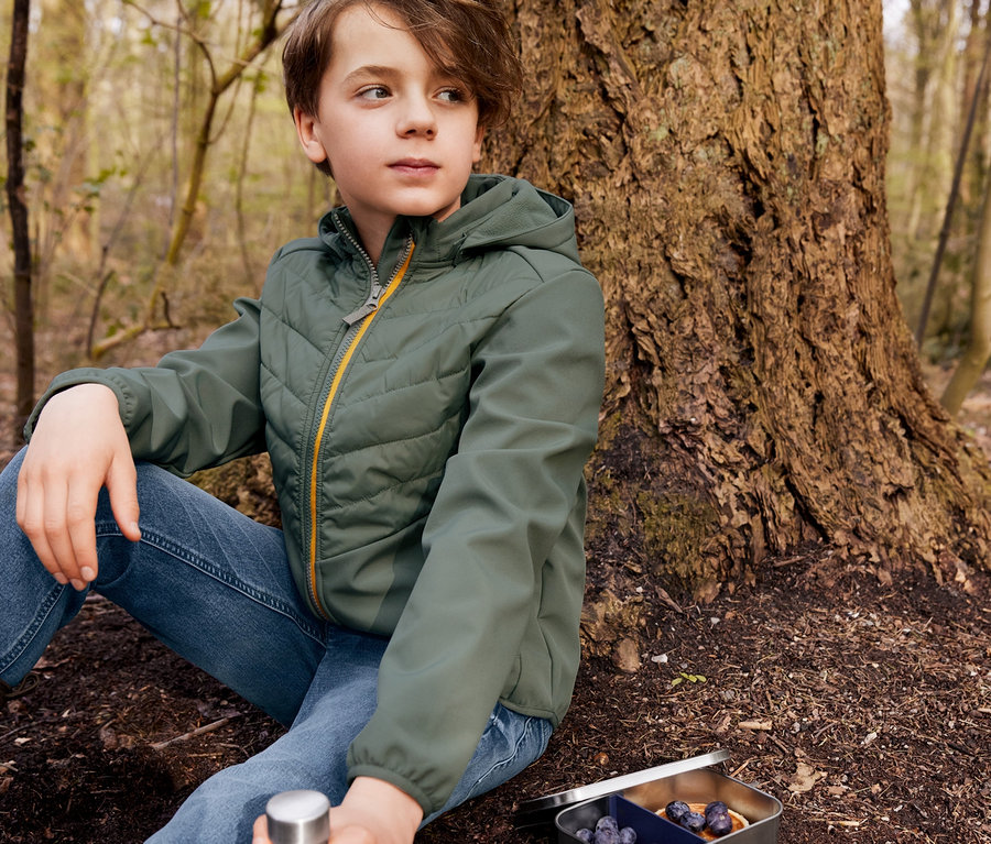 Ein Junge sitzt an einem Baum in blauen Jeans und einer Hybridjacke. Neben ihm liegen eine Brotdose und eine Trinkflasche.