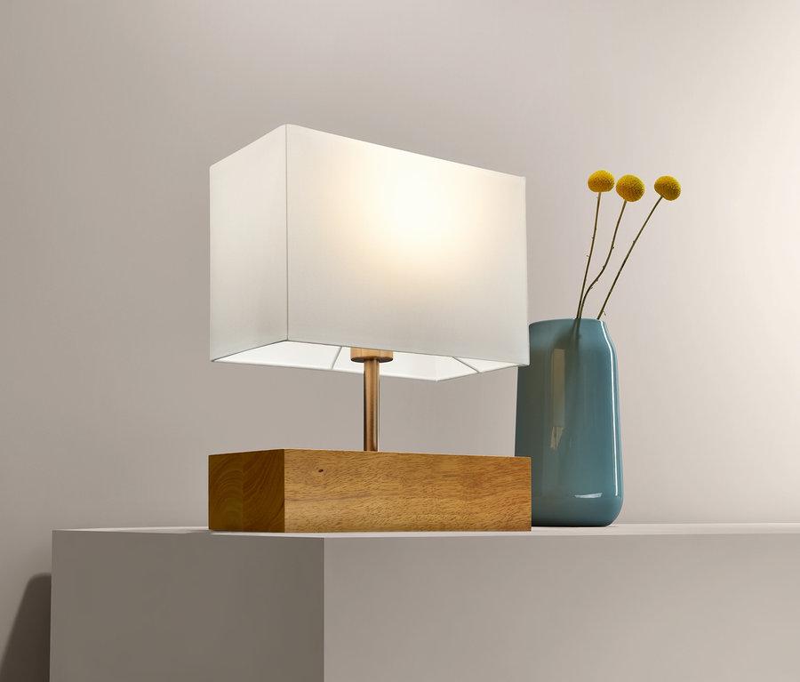 Eine Tischleuchte mit Holzfuß steht neben einer blauen Vase mit gelben Blumen auf einem weißen Regal.