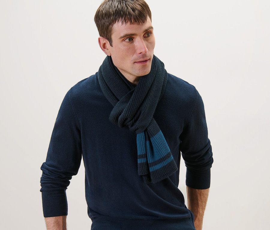 Mann mit Strickschal, marineblauem Pullover und Jeans, der sich an einen Stuhl lehnt.