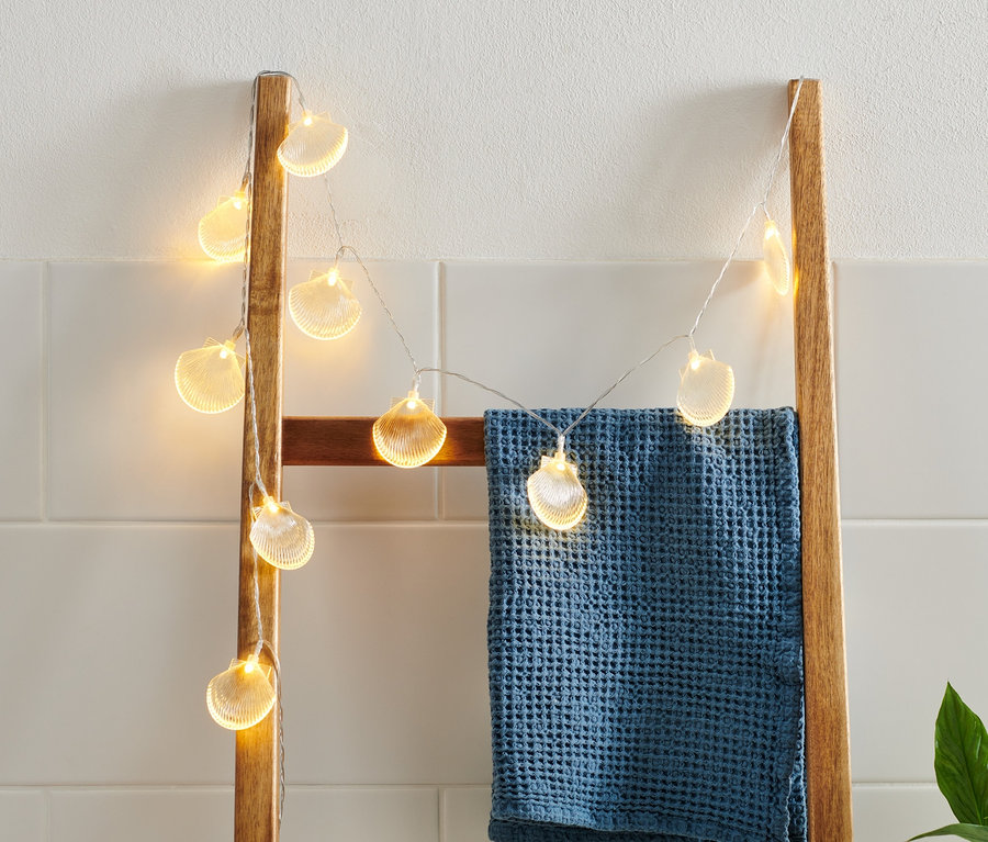 Eine LED-Lichterkette mit Muscheln hängt an einer Holzleiter, über die ein blaues Handtuch geworfen ist.
