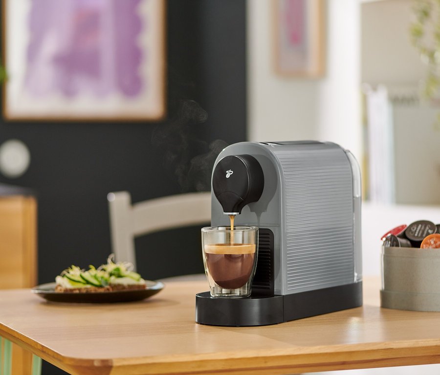 Eine graue Cafissimo pure+ Kaffeemaschine gießt Kaffee in ein Glas. Auf dem Tisch steht ein Teller mit belegten Broten.