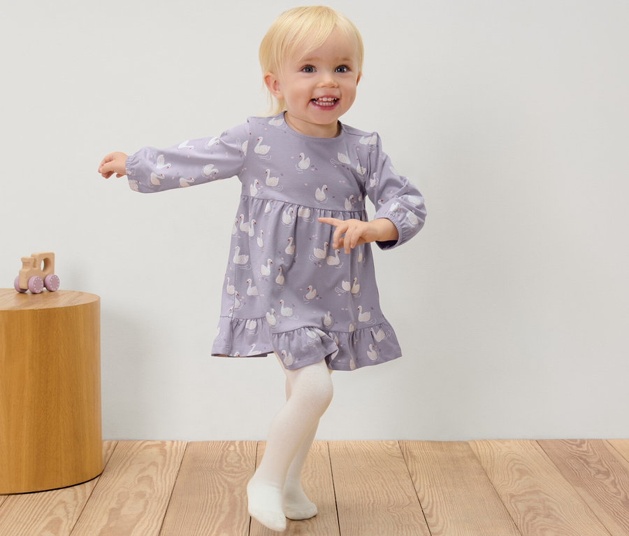 Ein Mädchen in einem Kleid mit Schwänen und Strumpfhosen steht neben einem Holztisch mit einem Spielzeug.