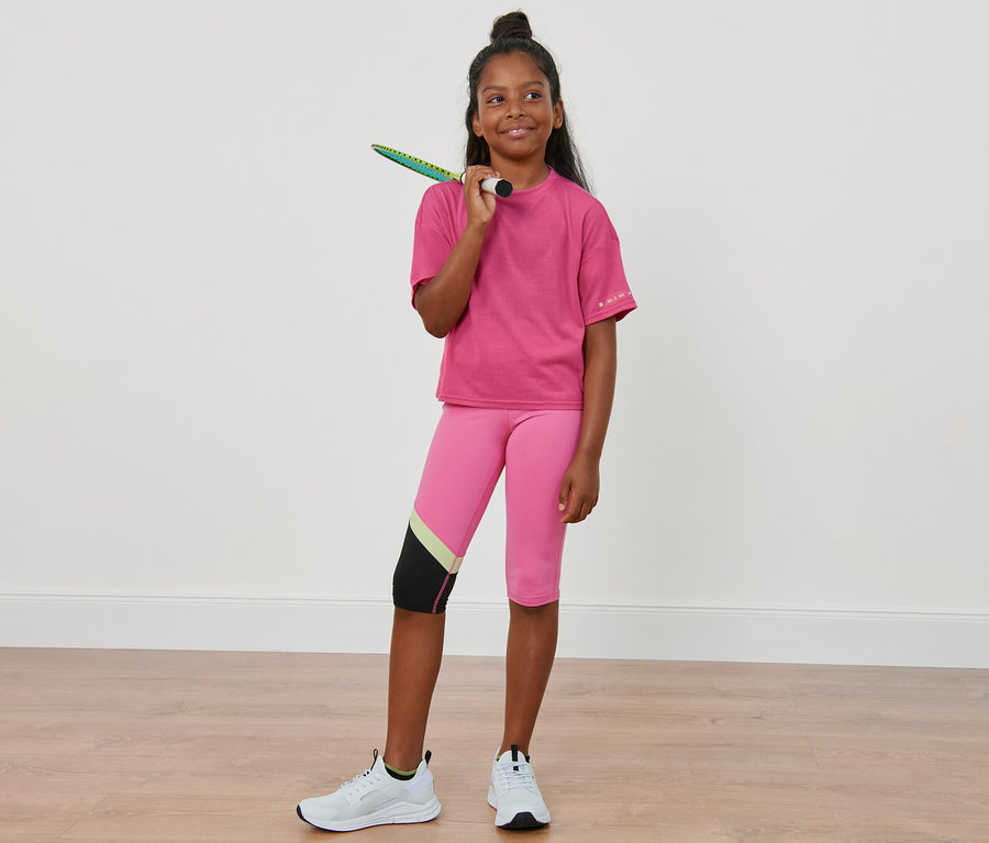 Mädchen mit Badmintonschläger über der Schulter trägt 2 Kinder-Sportshirts, rosa Leggings und weiße Turnschuhe.