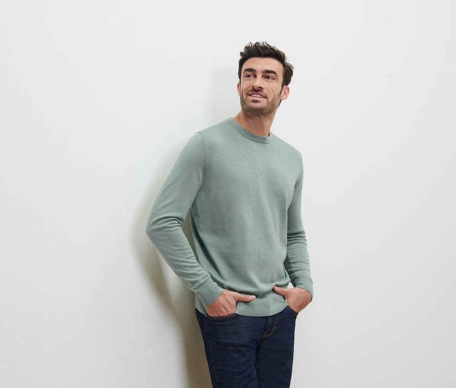Ein Mann posiert mit den Händen in den Hosentaschen vor einem hellen Hintergrund und trägt einen hellgrünen Merino-Pullover.