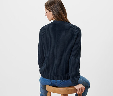 Frau sitzt auf einem Holzstuhl und trägt einen dunkelblauen Grobstrick-Cardigan.