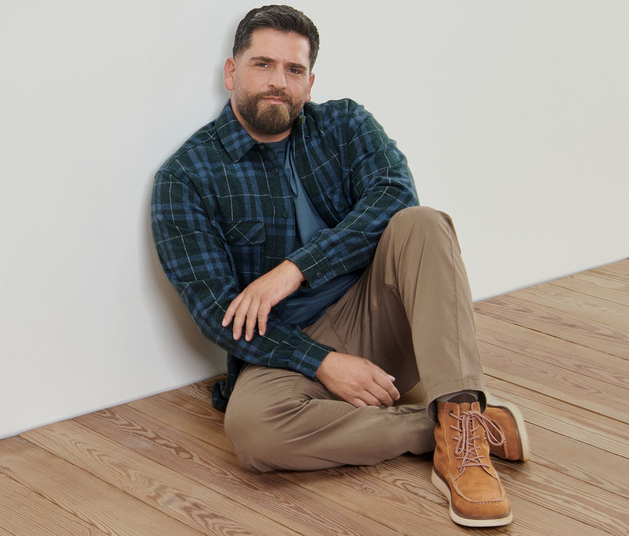 Ein Mann sitzt auf einem Holzboden und trägt eine sandfarbene Chino, eine Flanell-Hemdjacke und 2 Langarmshirts.