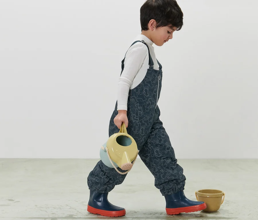 Junge geht und trägt eine Gießkanne. Er trägt eine dunkelblaue Kinder Thermo-Regenhose mit reflektierenden Elementen.