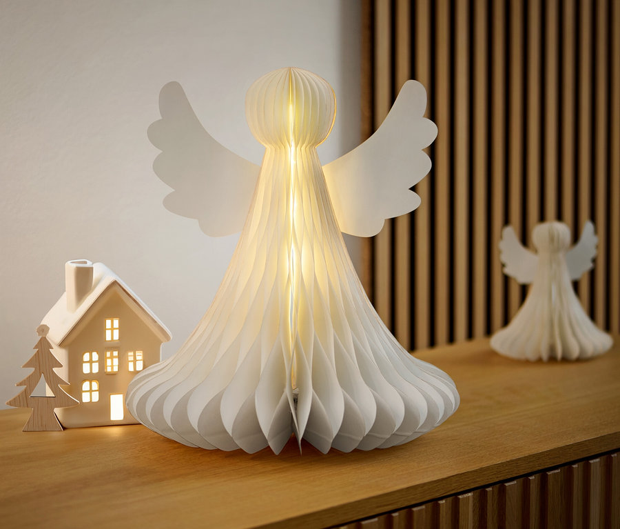 Ein großer LED-Papier-Engel steht auf einer Holzkommode neben einem beleuchteten Keramikhaus.