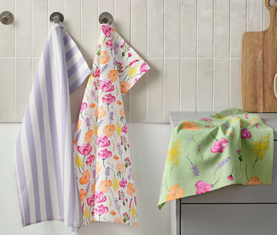 Drei Geschirrtücher hängen an der Wand: eines lila-weiß gestreift, eines mit Blumenmuster und eines liegt auf der Theke. Daneben befindet sich ein Schneidebrett und eine Tasse mit Küchenutensilien. 3 Geschirrtücher