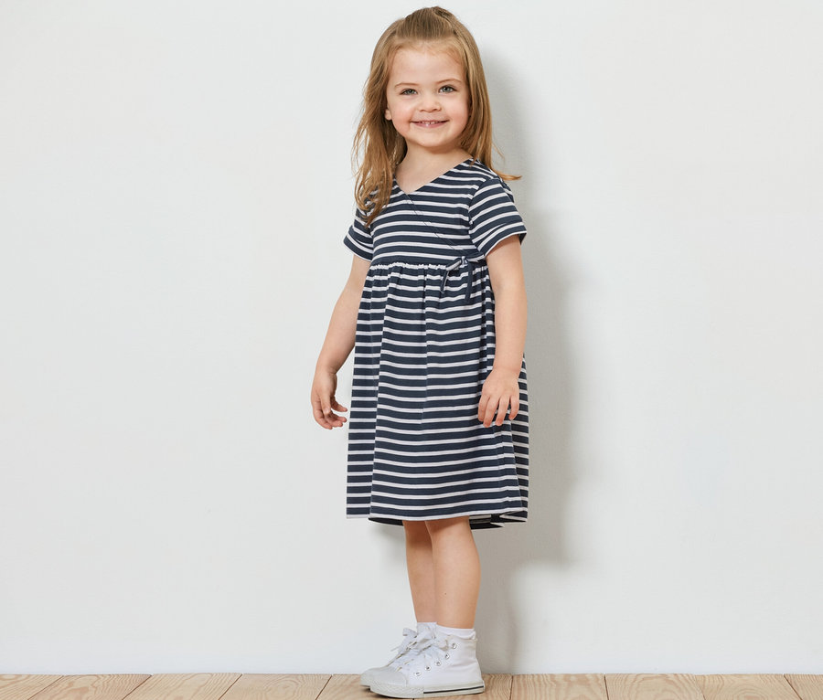 Ein lächelndes Mädchen steht seitlich und trägt ein Kinder-Jerseykleid mit blau-weißen Streifen und weiße Turnschuhe.