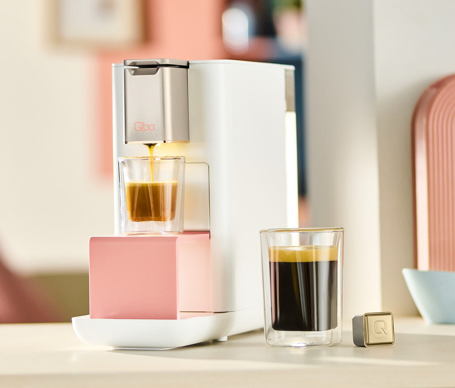 Qbo ICONIC x Pantone Special Edition Muted Clay Kaffeemaschine füllt ein Glas mit Kaffee. Daneben stehen eine Tasse Espresso und eine Kapsel.