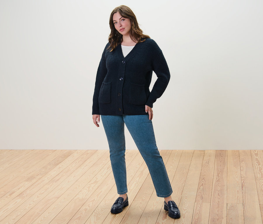 Eine Frau steht und trägt einen dunkelblauen, gerippten Grobstrick-Cardigan, ein weißes Shirt mit 3/4-Arm und eine Straight Jeans – Fit »Juna«.