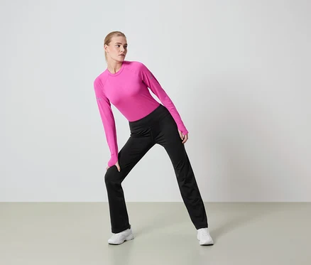 Frau mit blonden Haaren dehnt sich in einem pinkfarbenen Langarmshirt und einer schwarzen Hose.