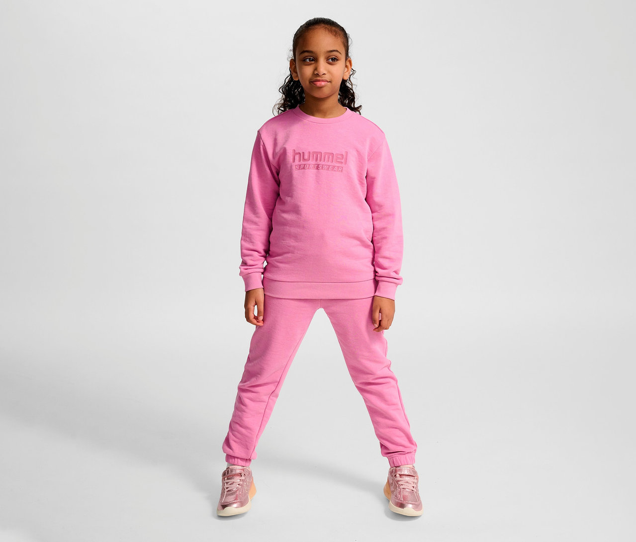 Junges Mädchen posiert in einem rosa HUMMEL HMLJR Base-Sweatshirt und Jogginghose.