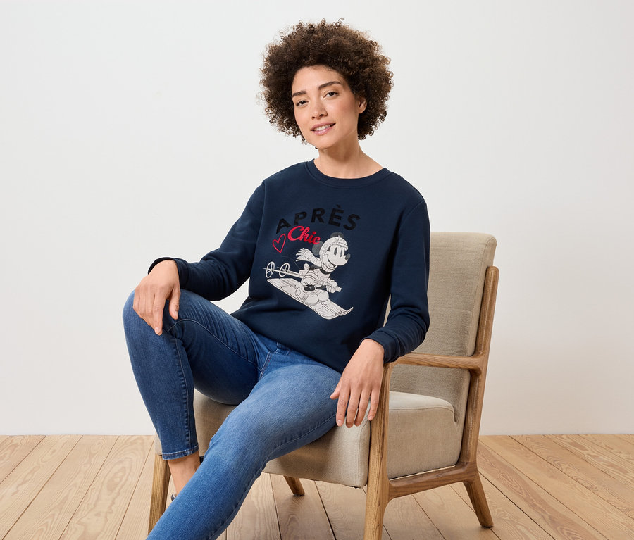 Eine Frau sitzt in einem Sessel und trägt einen marineblauen Pullover mit einem Bild von Mickey Mouse auf Skiern und blaue Jeans.
