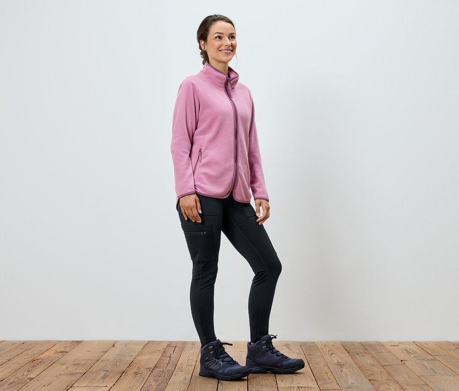 Frau in rosa Fleecepullover, schwarzen Hosen und Stiefeln steht auf Holzboden.