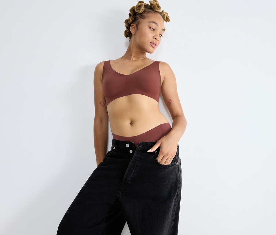 Eine junge Frau mit Duttfrisur lehnt an einer weißen Wand und trägt einen braunen sloggi ZERO Feel 2.0 Bralette und passende Unterwäsche unter einer offenen schwarzen Jeans.