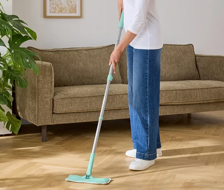 Frau wischt einen Holzboden mit einem Mopp mit blaugrüner Unterlage.