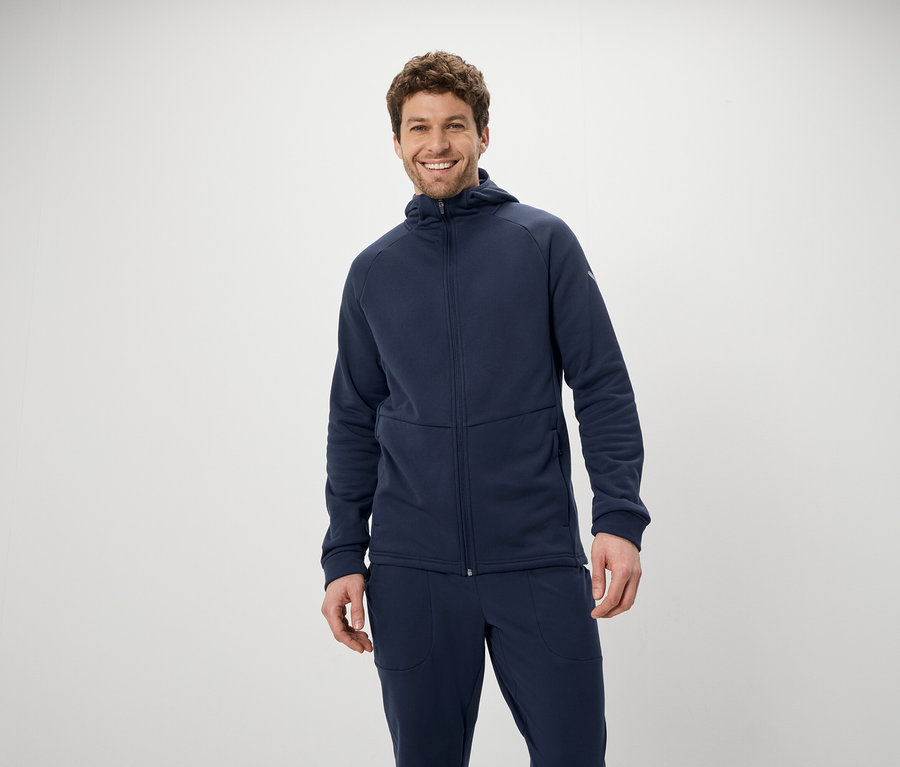 Ein lächelnder Mann posiert in einem blauen Trainingsanzug mit Kapuzenjacke und Hose vor einem weißen Hintergrund.