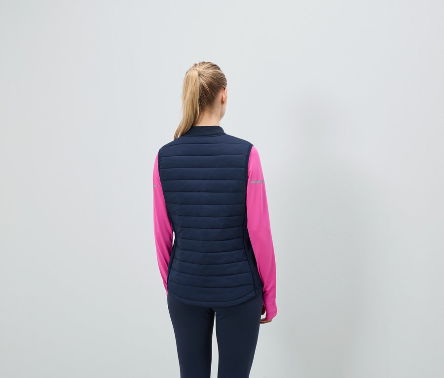 Rückansicht: Frau mit blonden Haaren zum Pferdeschwanz, trägt eine dunkelblaue Steppweste und ein pinkes Sporttrikot.