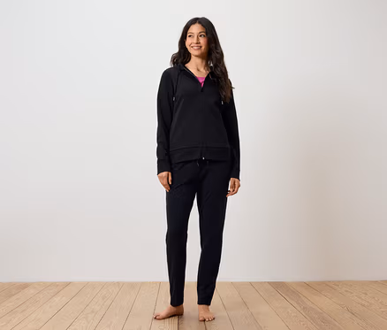 Eine lächelnde Frau steht barfuß auf einem Holzboden. Sie trägt eine schwarze Hose und einen schwarzen Kapuzenpullover mit Reißverschluss.