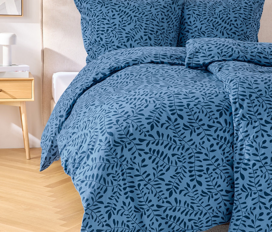 Ein blaues Bettwäsche-Set mit Blattmuster auf einem Bett mit Kopfteil. Ein Nachttisch mit Lampe steht neben dem Bett.