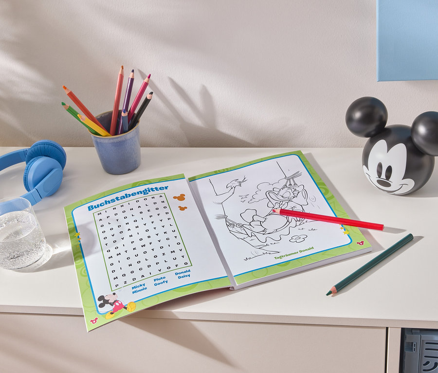 Auf einem weißen Tisch liegen ein aufgeschlagenes Malbuch, bunte Stifte in einem blauen Becher, blaue Kopfhörer, ein Glas Wasser und eine Mickey Mouse-Figur.