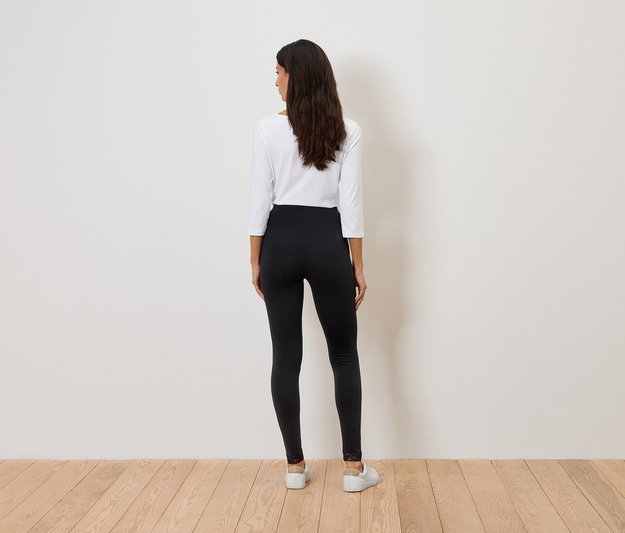 Rückansicht einer Frau mit langen, dunklen Haaren, die ein weißes Oberteil und schwarze Leggings mit Spitzenbesatz trägt.