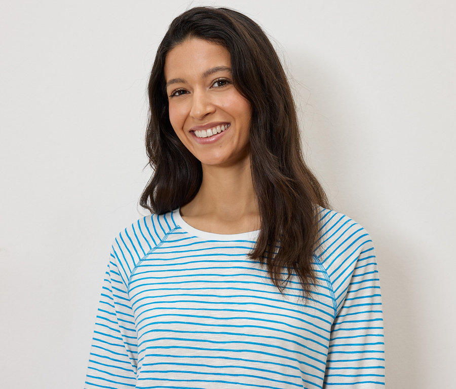 Frau lächelt und trägt ein blau-weiß gestreiftes Langarmshirt vor einem weißen Hintergrund.