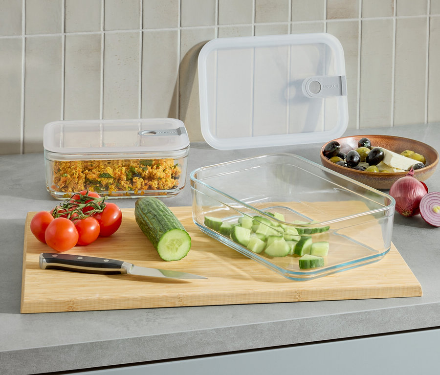 Auf einer Küchentheke stehen zwei Glasbehälter, einer mit Essen und einer mit geschnittenen Gurken. Außerdem gibt es Tomaten, eine Gurke, ein Messer und eine Schale mit Oliven und Käse.