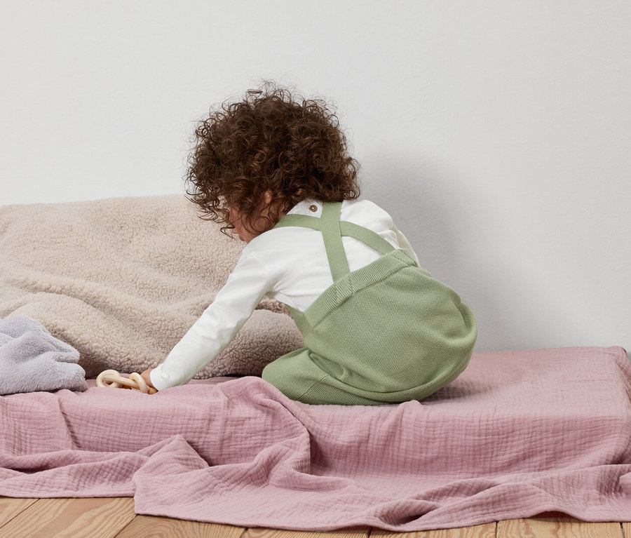 Kind mit lockigem Haar kniet auf einer rosa Decke und trägt eine grüne Latzhose und ein langärmliges Hemd.