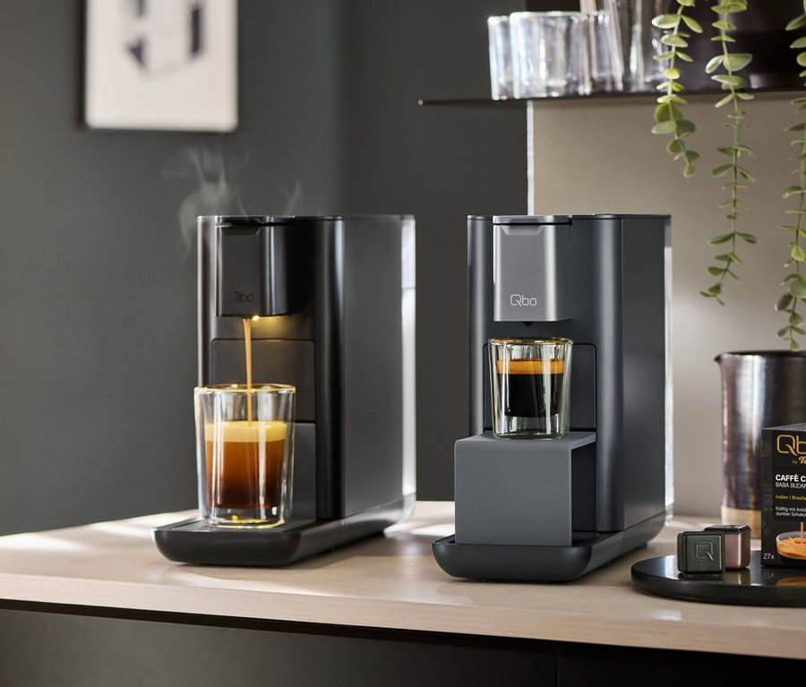 Zwei Kaffeemaschinen auf einer Arbeitsplatte. Die linke Qbo ICONIC Dark Chrome brüht Kaffee in ein Glas, während die rechte Qbo ESSENTIAL Dark Stone ein Glas mit Kaffee bereithält. Daneben stehen Qbo by Tchibo Caffè Crema Kapseln.