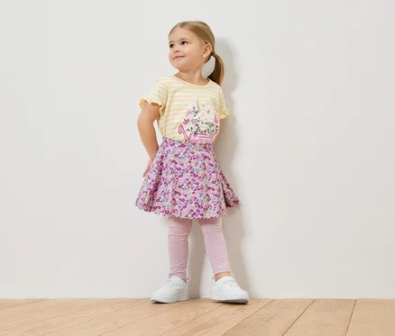 Ein kleines Mädchen mit blonden Haaren trägt ein gelb-gestreiftes Shirt mit einem Blumenmuster, einen geblümten Rock, rosa Leggings und weiße Turnschuhe.