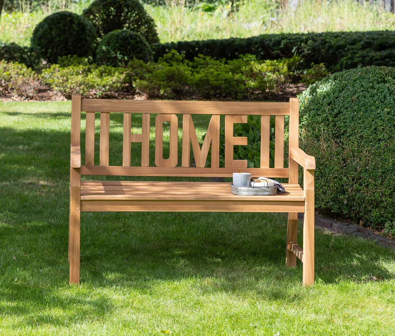 Eine deVries Gartenbank »Home« steht auf einer Wiese vor Büschen.