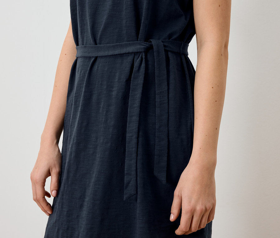 Nahaufnahme einer Frau, die ein ärmelloses, marineblaues Kleid mit Stoffgürtel trägt.