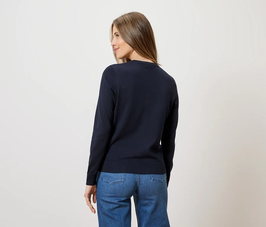 Eine Frau mit langen braunen Haaren trägt einen dunkelblauen Pullover und Jeans.