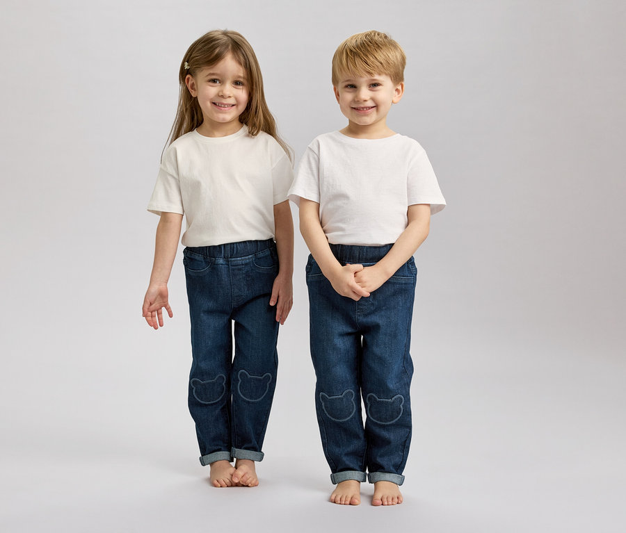 Zwei Kinder mit blonden und braunen Haaren stehen barfuß nebeneinander und tragen weiße T-Shirts und blaue Jeans mit Bärenmotiven.