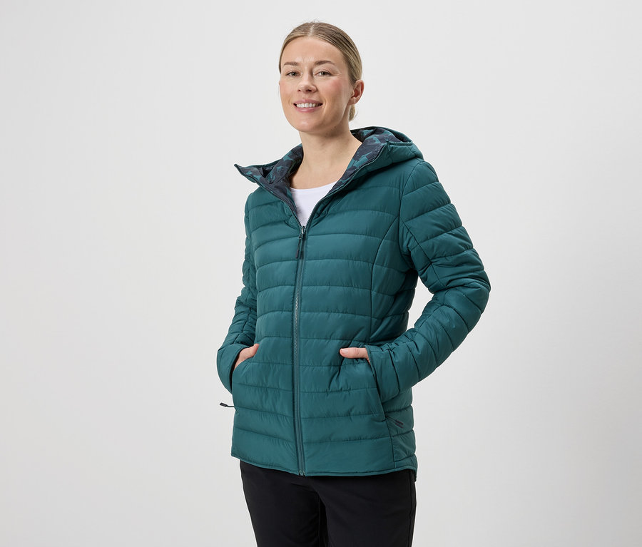 Frau mit Kapuze trägt eine smaragdgrüne Steppjacke mit Reißverschluss und schwarzen Hosen vor einem neutralen Hintergrund.