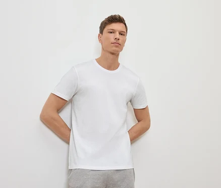 Mann lehnt an einer weißen Wand und trägt ein weißes T-Shirt und graue Jogginghose.