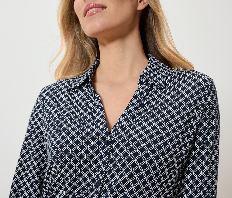 Oberkörper einer Frau, die eine dunkelblaue Bluse mit weißem geometrischem Muster trägt.